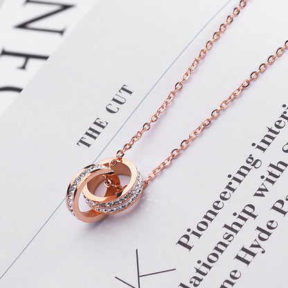 Rose gold double-ring pendant necklace with small clear stones, displayed on a white printed paper background with partial text visible.