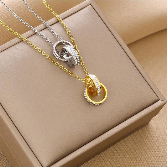 Two interlocking ring-style necklaces displayed on a beige jewelry box. One necklace is gold with a clear stone-set design, and the other is silver with a clear stone-set design, both on delicate chains.